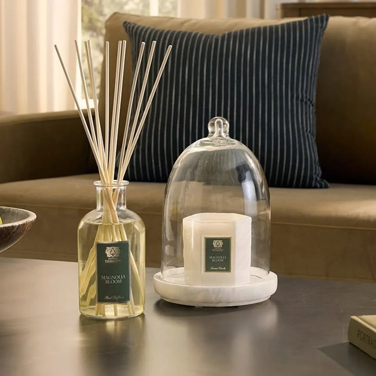 A reed diffuser and candle under a glass cloche on a table with a sofa and striped pillow in the background.
