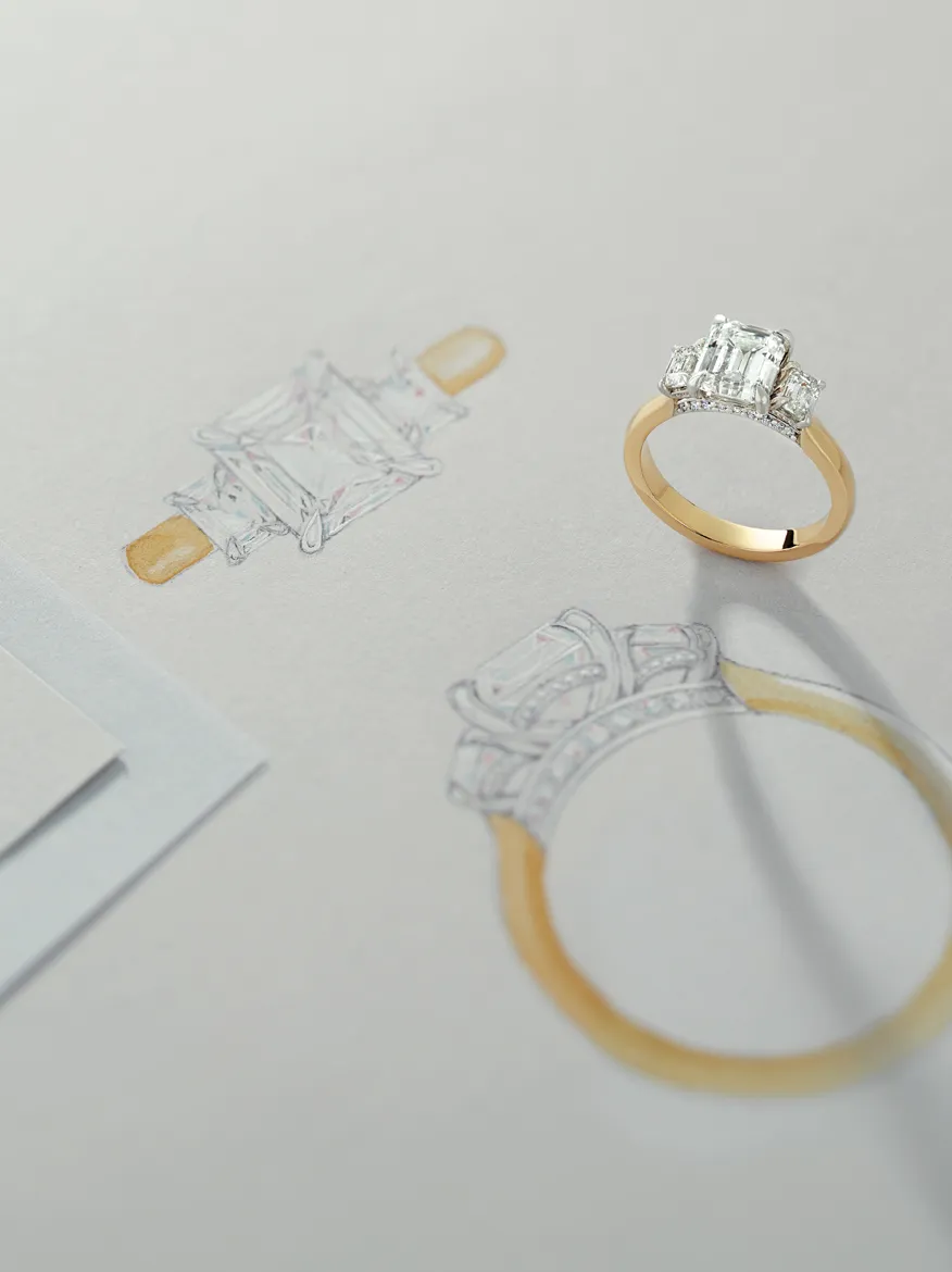 An elegant gold engagement ring beside a detailed sketch of the same design on paper.