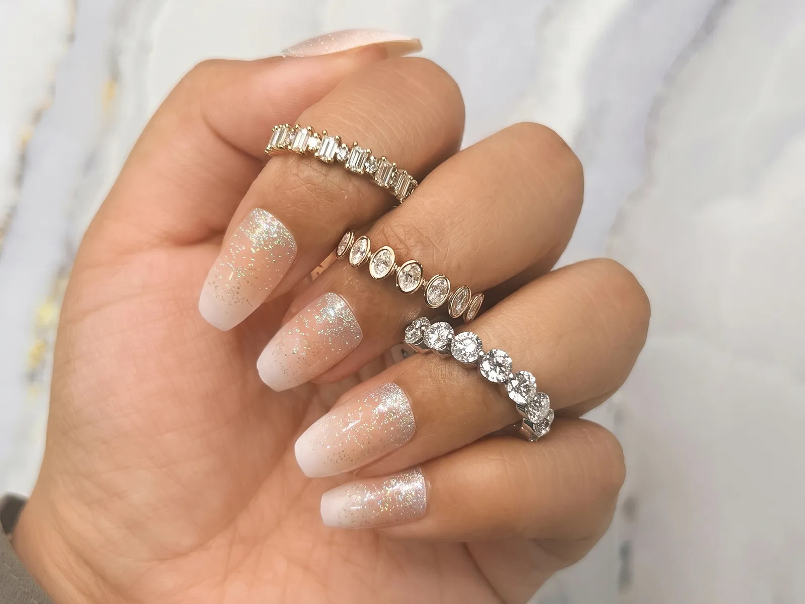 A hand with glittery ombre nails wearing three diamond rings on different fingers.