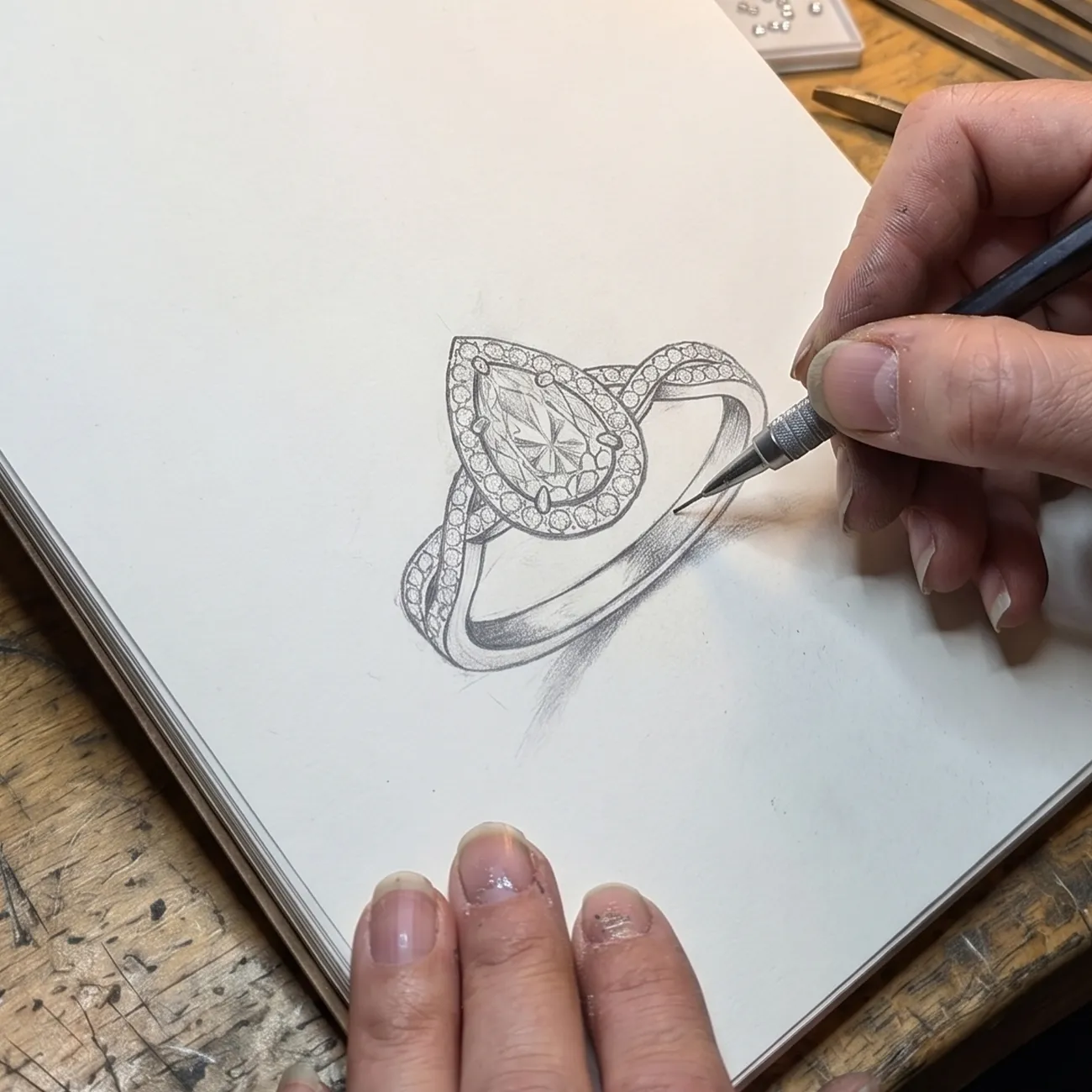 A hand sketching a detailed diamond ring design on paper with a pencil.
