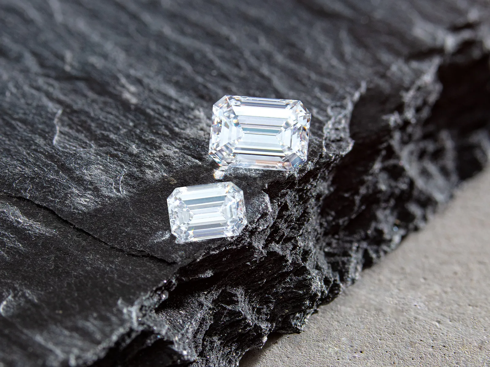 Two emerald-cut diamonds on a textured dark stone surface.