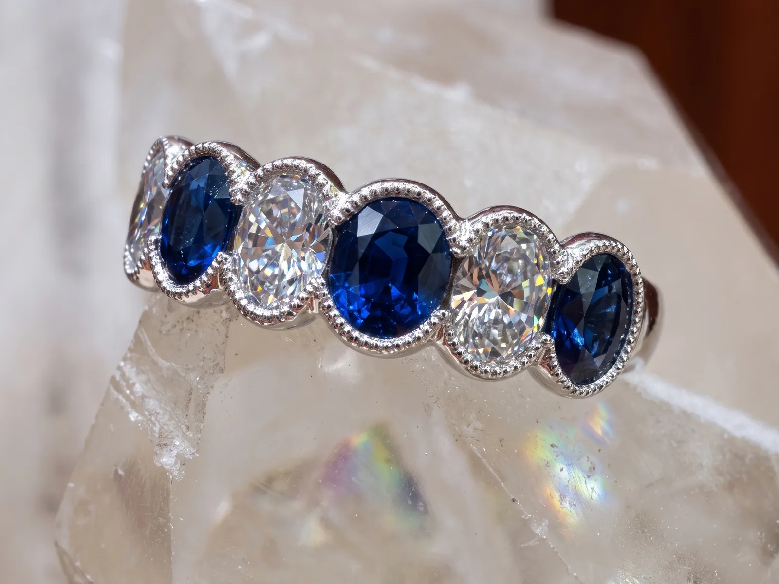 A ring with alternating blue sapphires and diamonds set in silver on a crystal surface.