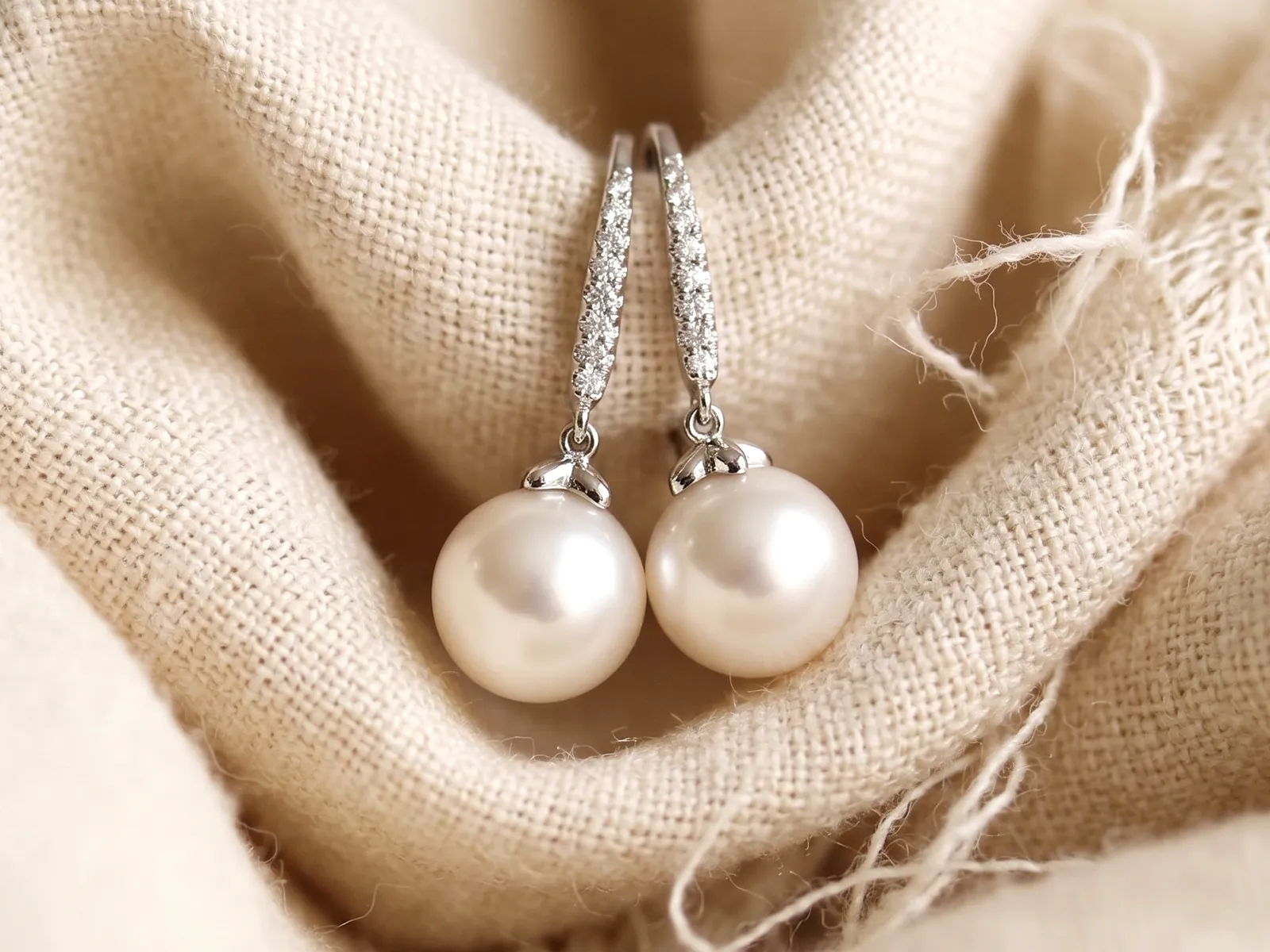Elegant pearl and diamond earrings on textured beige fabric background.