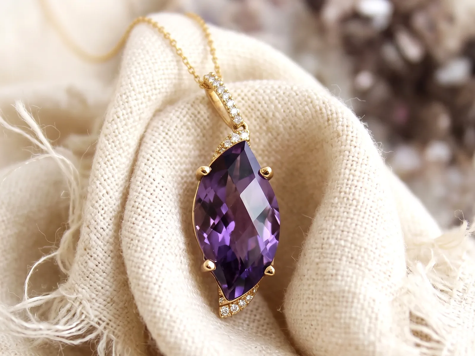 A marquise-cut purple gemstone pendant with diamonds on a gold chain, displayed on a textured fabric.