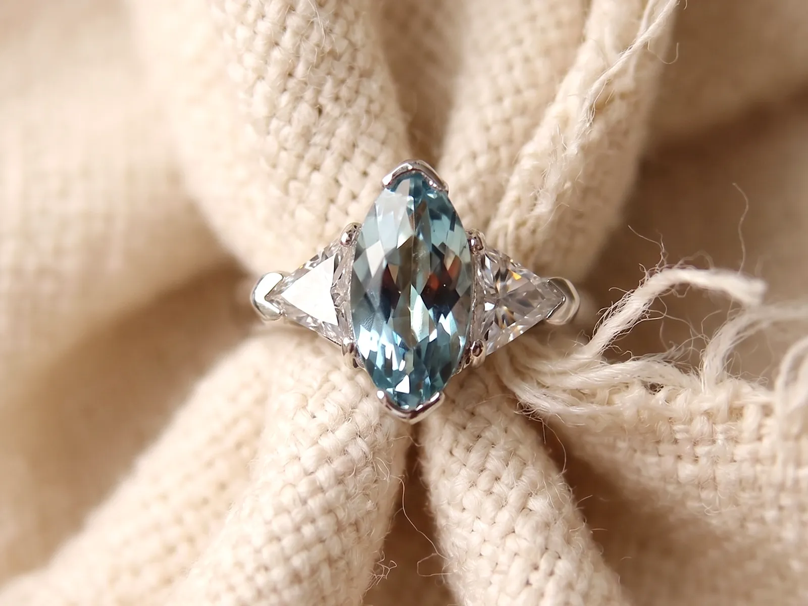 A marquise-cut blue gemstone ring with side stones on a textured fabric background.