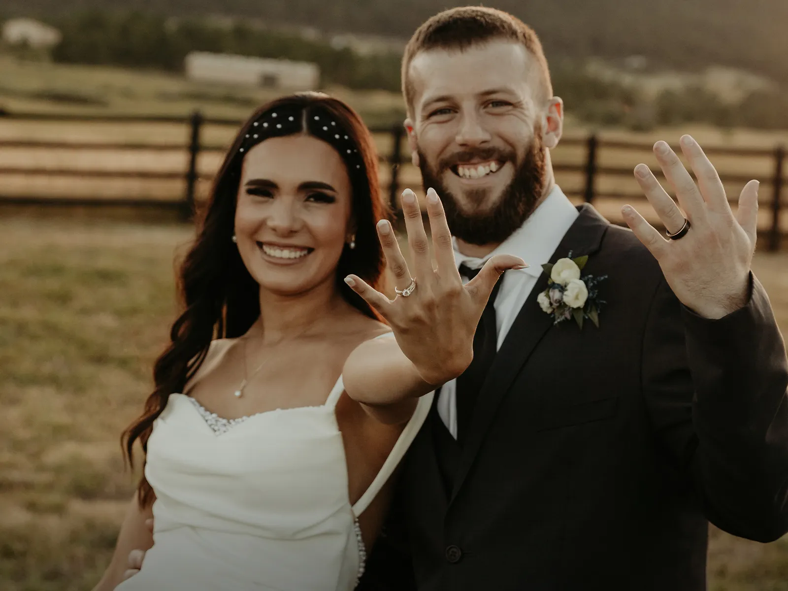 A smiling couple in wedding attire shows off their rings in a scenic outdoor setting.