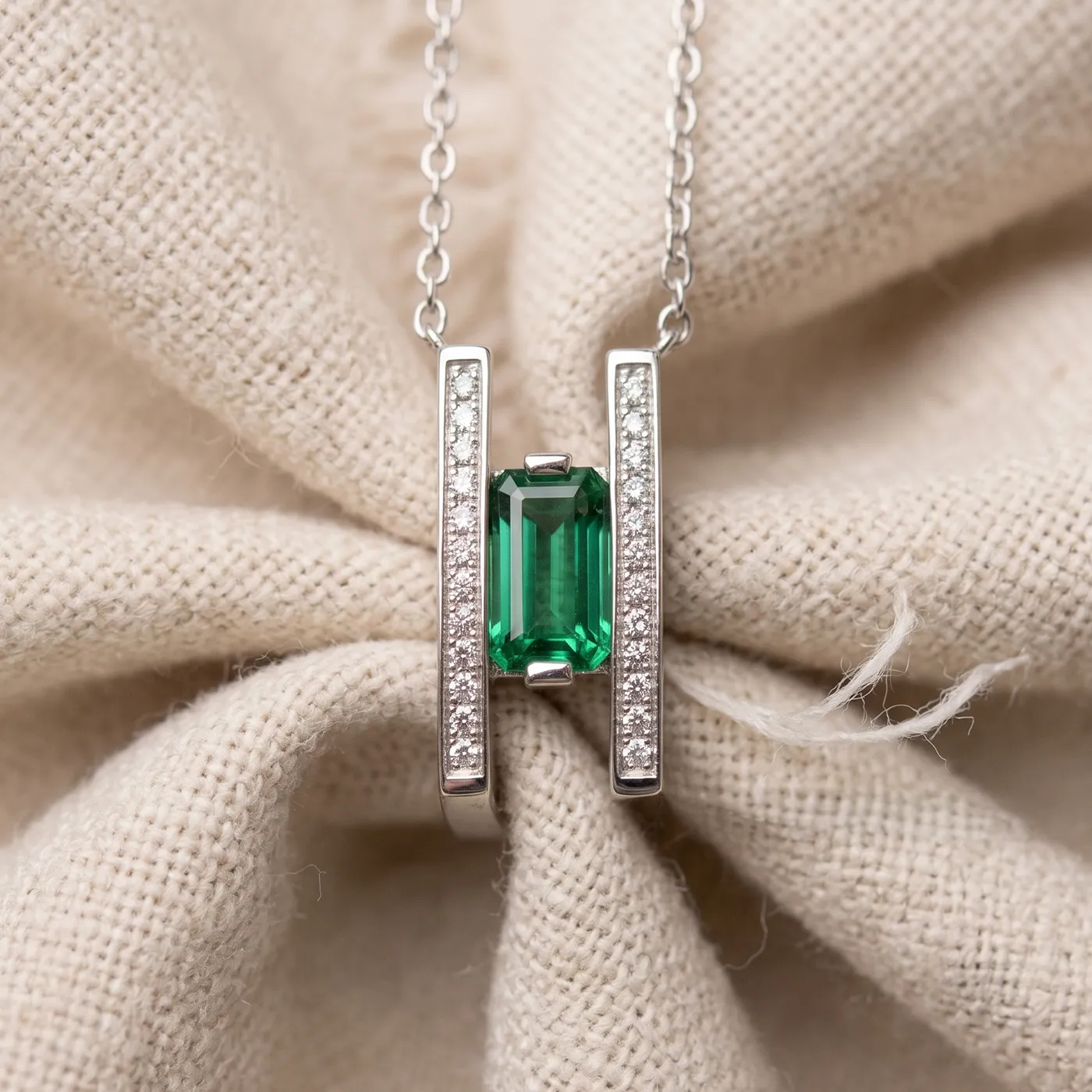 A silver necklace with a green gemstone pendant on a textured beige fabric background.