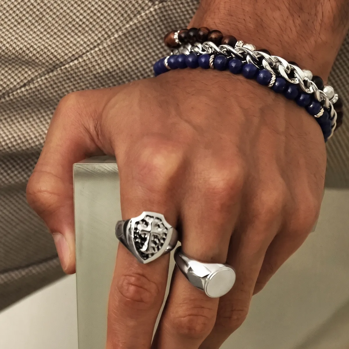 A hand wearing silver rings and multiple bracelets, including beaded and chain styles, on a textured surface.