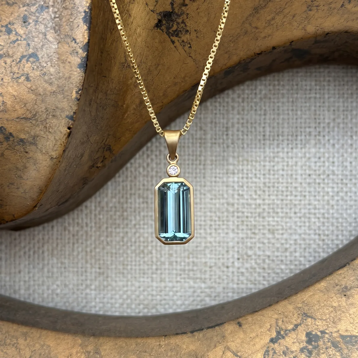 A gold necklace with a rectangular blue gemstone pendant and a small diamond accent on a textured background.
