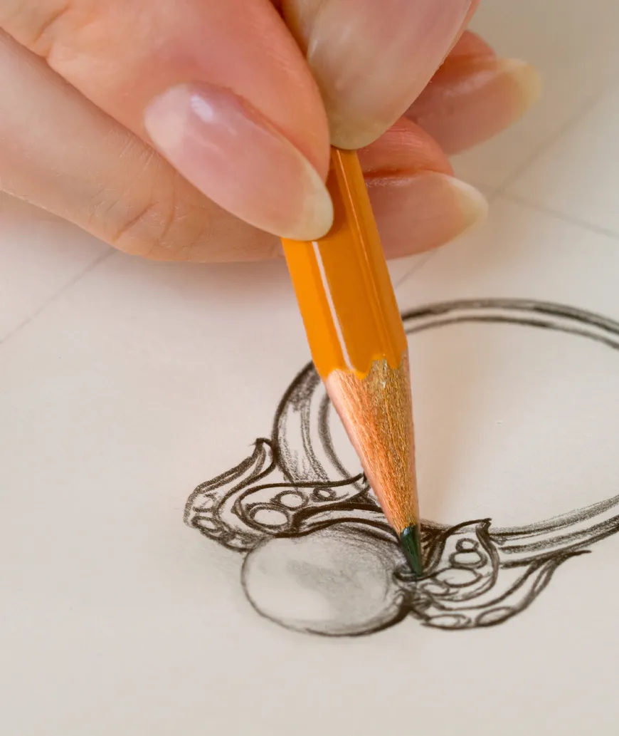 A hand sketching an intricate ring design with a pencil on paper.