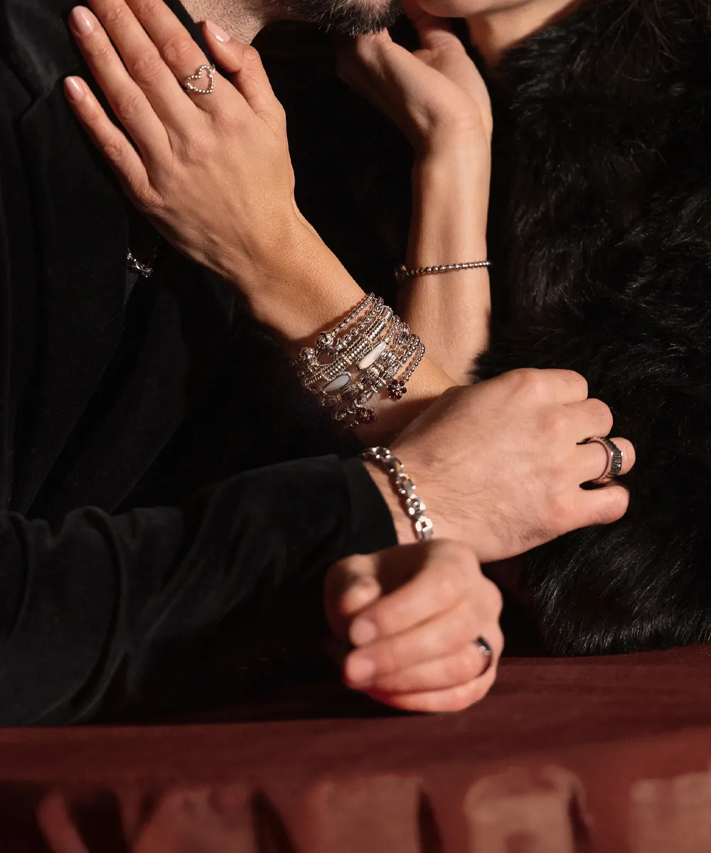 Two people with intertwined arms, showcasing bracelets and rings, in an intimate setting.