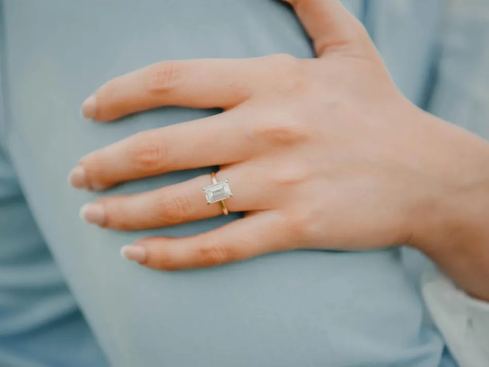 A hand with a rectangular diamond ring rests on a light blue fabric background.