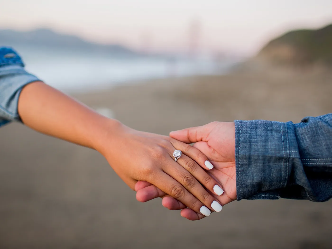 Hands holding on a beach, one with an engagement ring.