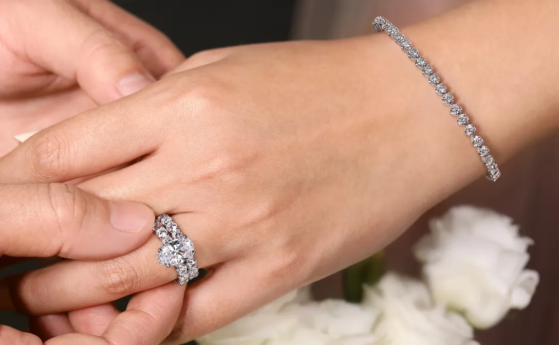 A hand wearing a diamond engagement ring and bracelet, with another hand gently holding it, surrounded by white flowers.