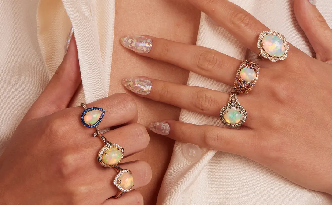 Five elegant rings featuring opals and colorful stones, displayed on manicured hands with shimmering nail art.