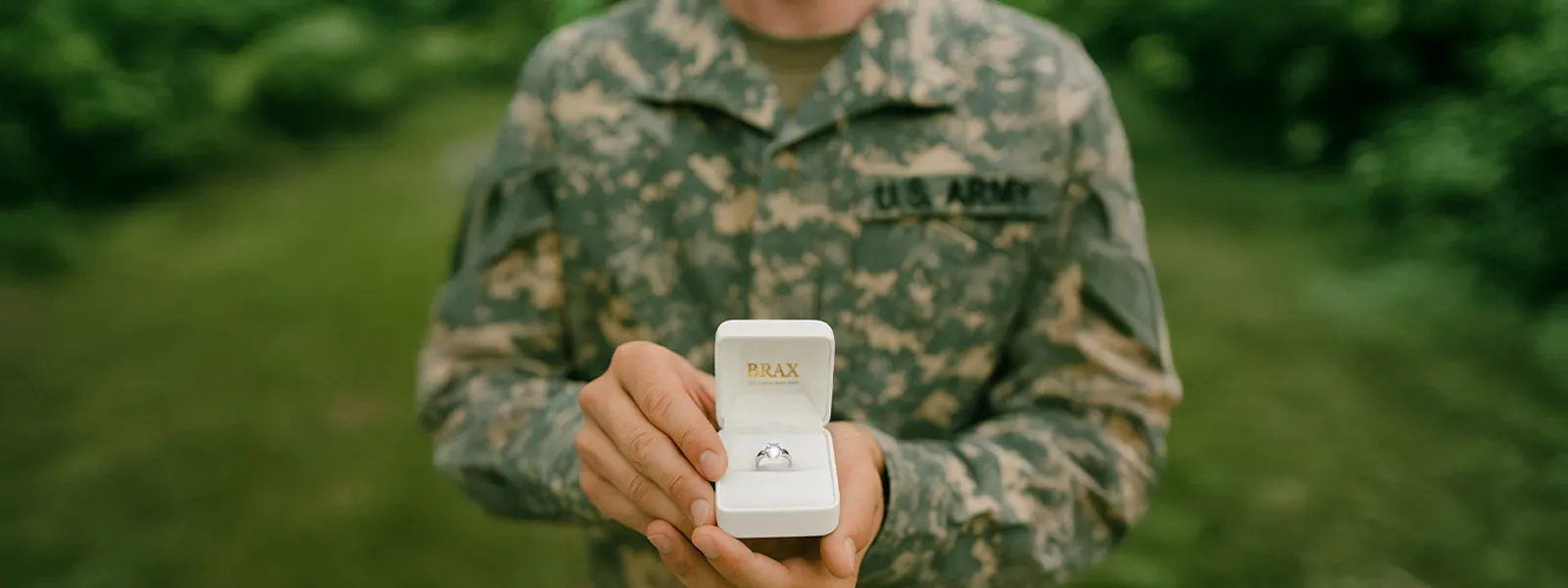 A US soldier and a woman holding hands, showcasing an engagement ring against a scenic backdrop.