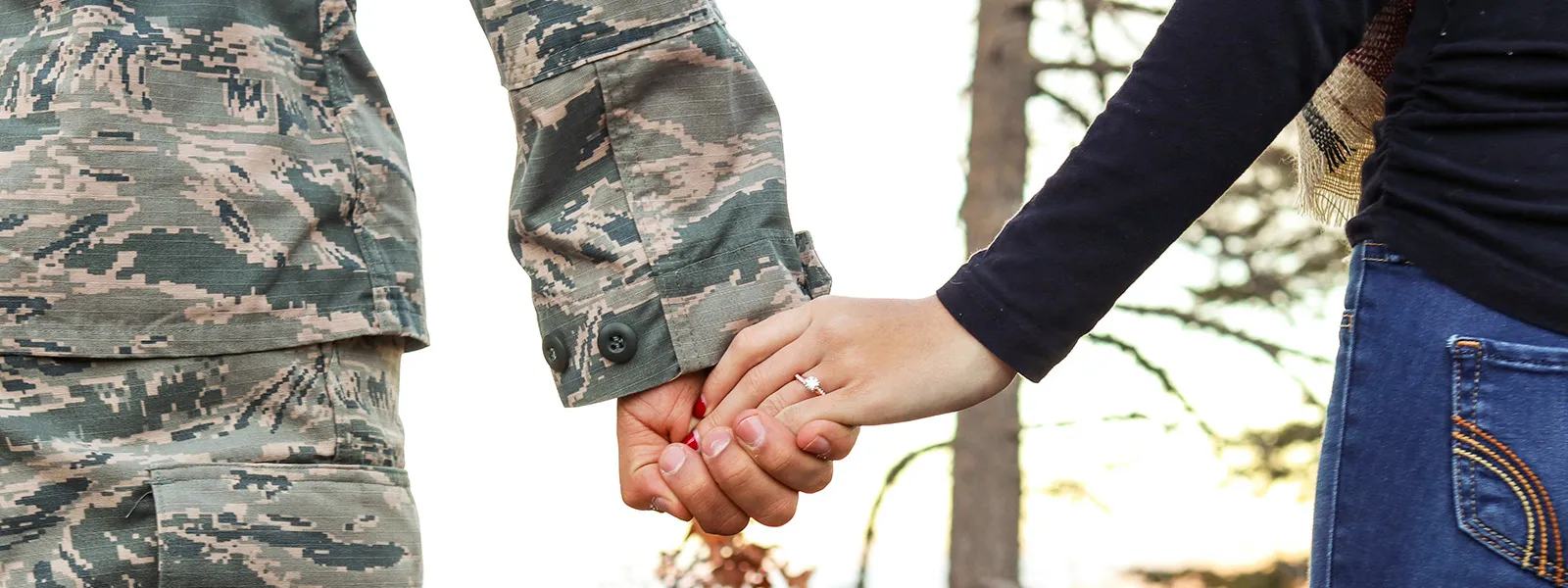 A US soldier and a woman holding hands, showcasing an engagement ring against a scenic backdrop.