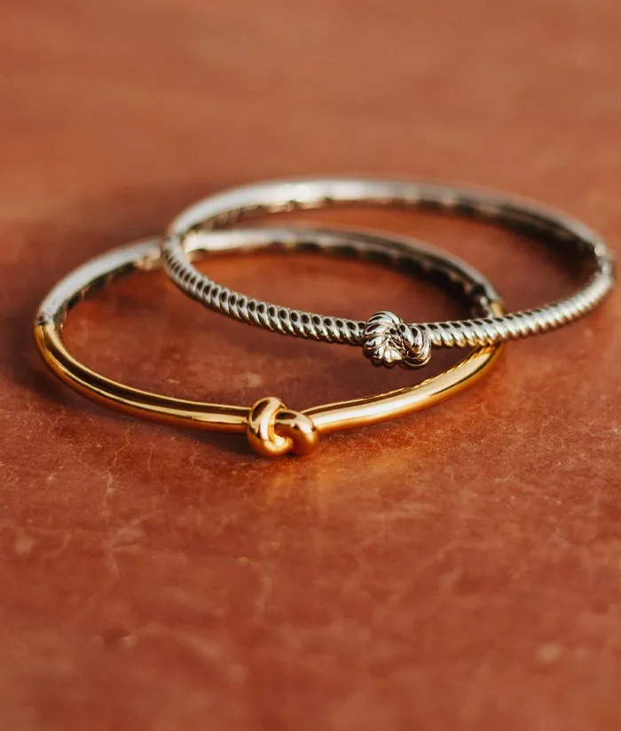 Three elegant bracelets: a gold bangle, a twisted silver design, and a silver knot bracelet on a textured background.