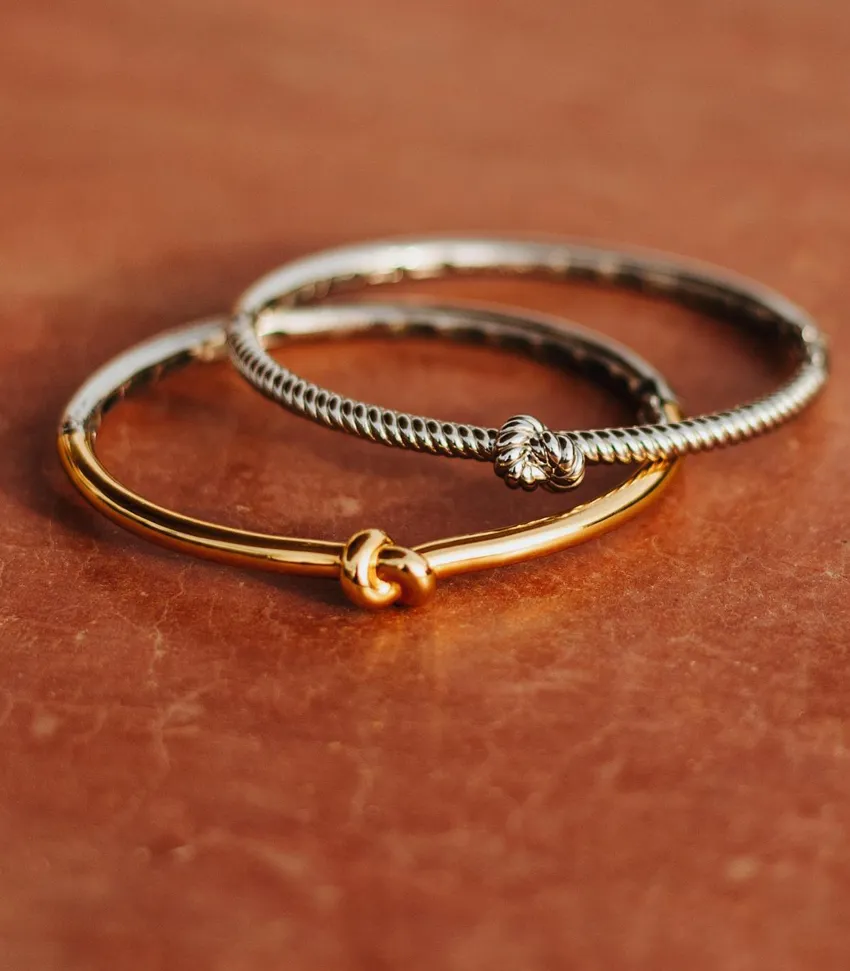 Three elegant bracelets: a gold bangle, a twisted silver design, and a silver knot bracelet on a textured background.