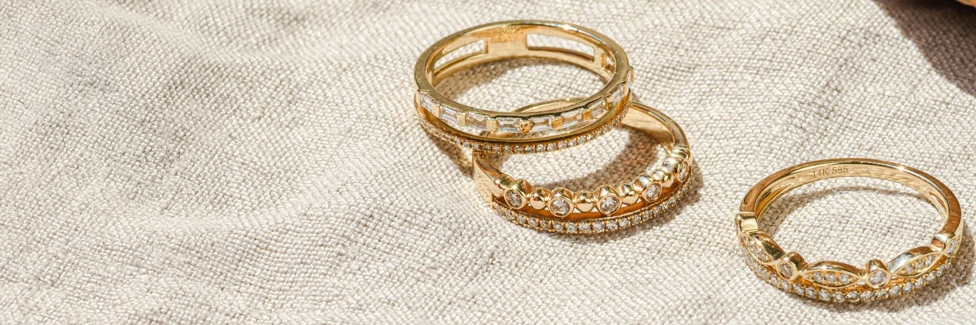 Three elegant gold rings with diamonds on a textured fabric background. Click to learn more about Bryan Jewelry's offerings.