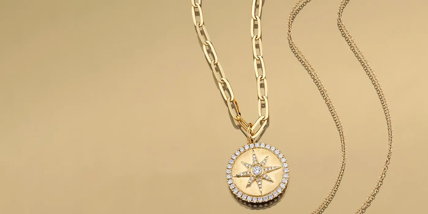 Gold compass pendant necklace with diamonds on a gold chain, displayed on a beige background. Shop at Bryan Jewelry in Prattvill