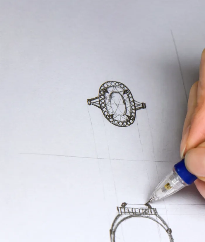 A hand sketches intricate ring designs on paper, showcasing custom jewelry creation for Bryan Jewelry in Prattville.