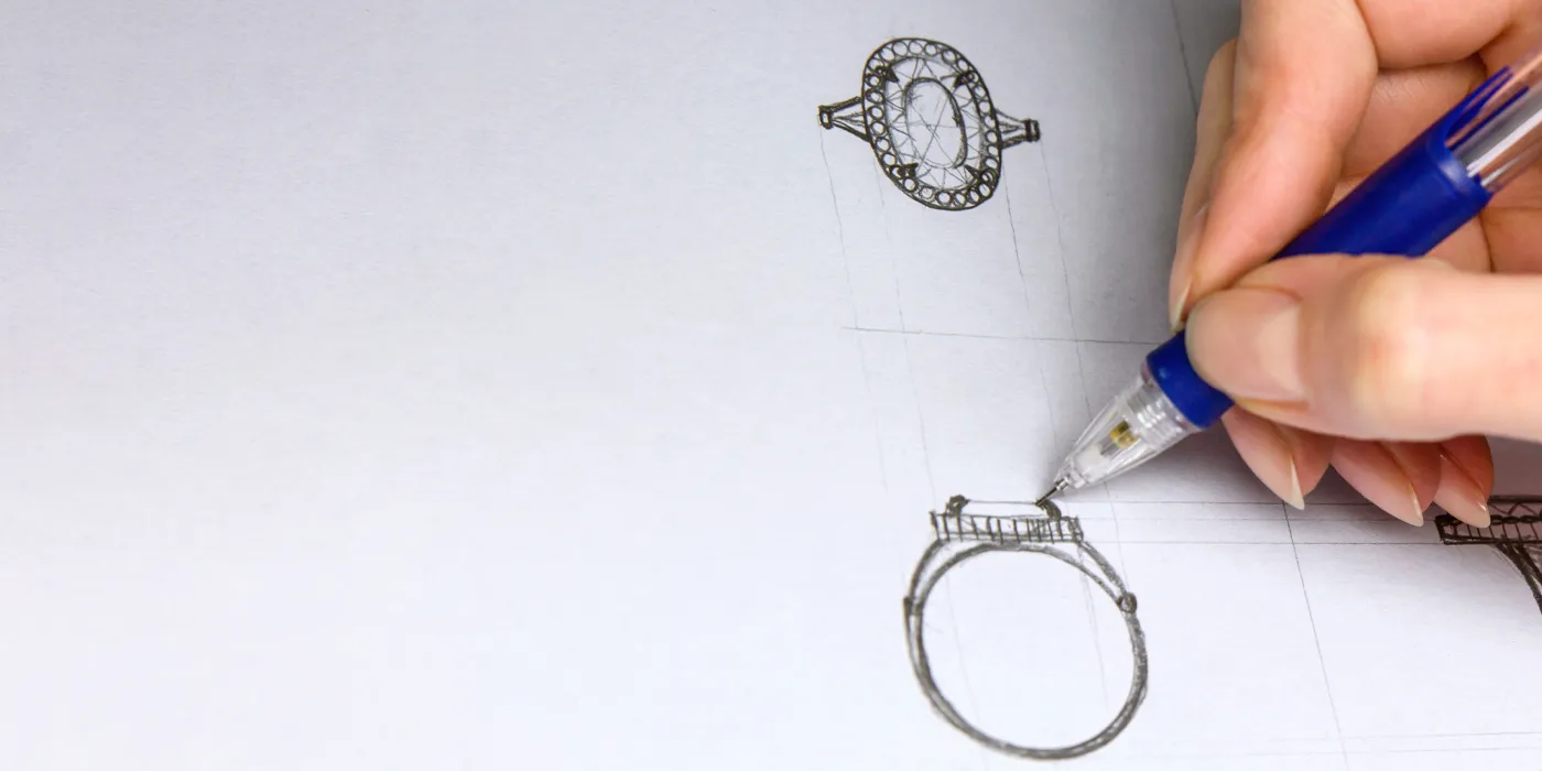 A hand sketches intricate ring designs on paper, showcasing custom jewelry creation for Bryan Jewelry in Prattville.