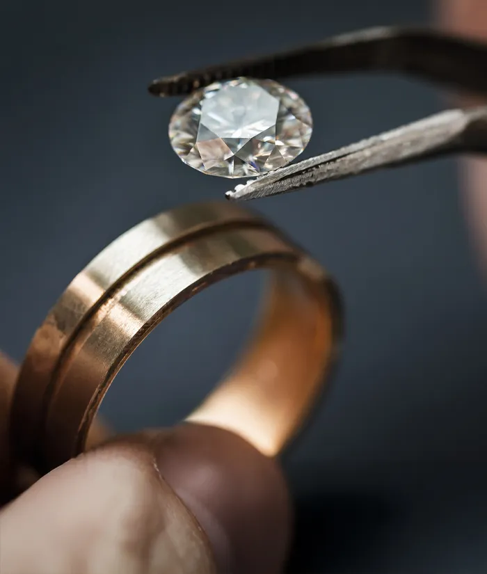 A jeweler using tweezers to hold a diamond above a gold wedding band, showcasing fine jewelry craftsmanship. Book an Appointment