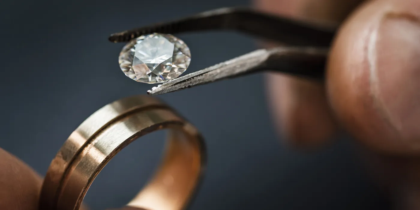A jeweler using tweezers to hold a diamond above a gold wedding band, showcasing fine jewelry craftsmanship. Book an Appointment