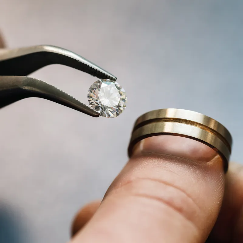 A jeweler using pliers to hold a diamond above a gold wedding band. Visit Bryan Jewelry for custom engagement rings.