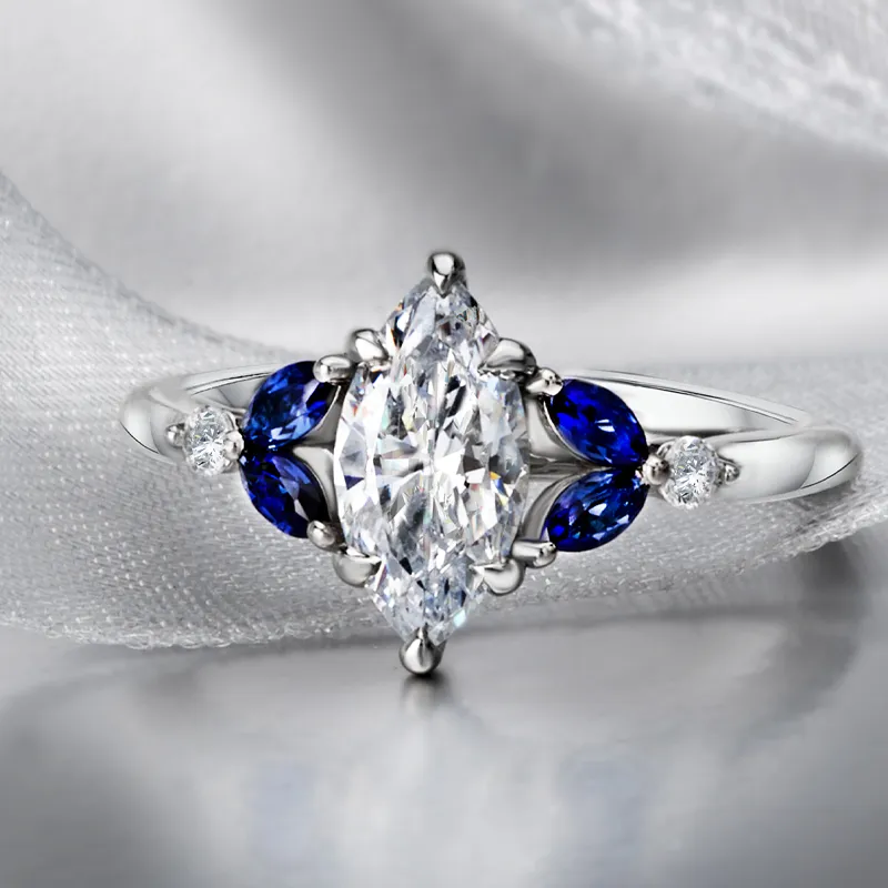 Elegant marquise diamond ring with blue sapphires and smaller diamonds, perfect for engagement. Visit Bryan Jewelry.
