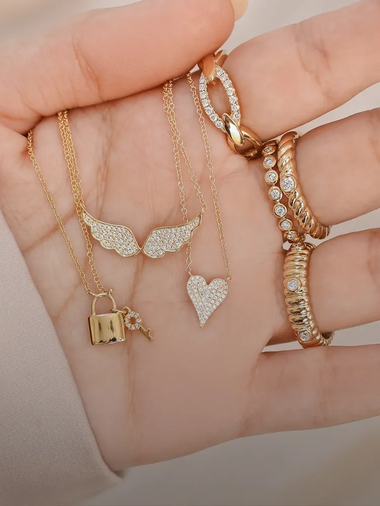 Gold necklaces and rings featuring charms like wings, a heart, and a padlock, displayed on a hand. 