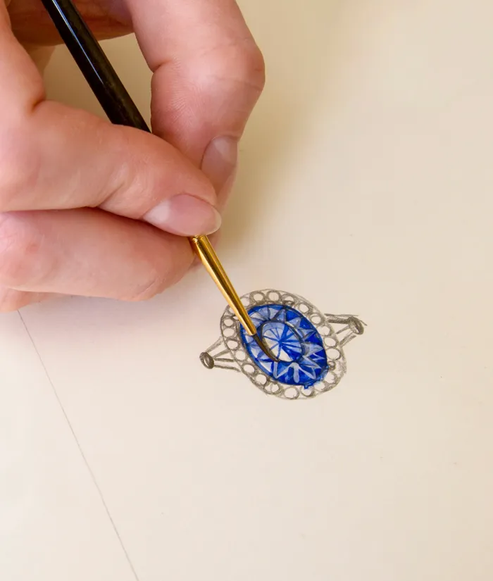 A hand painting a blue gemstone ring design on paper, showcasing custom jewelry creation at Bryan Jewelry.