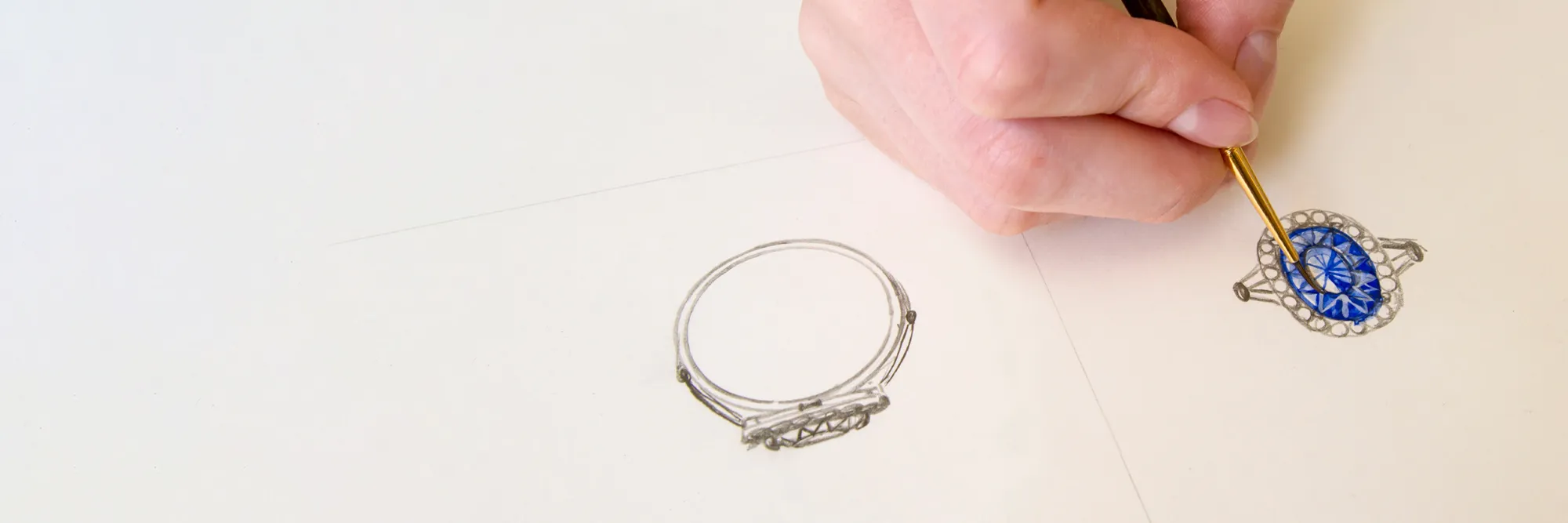 A hand painting a blue gemstone ring design on paper, showcasing custom jewelry creation at Bryan Jewelry.