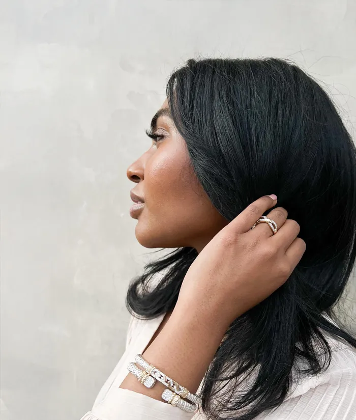 A woman with long black hair showcases elegant jewelry, including a bracelet and ring, against a light background. Shop Designer