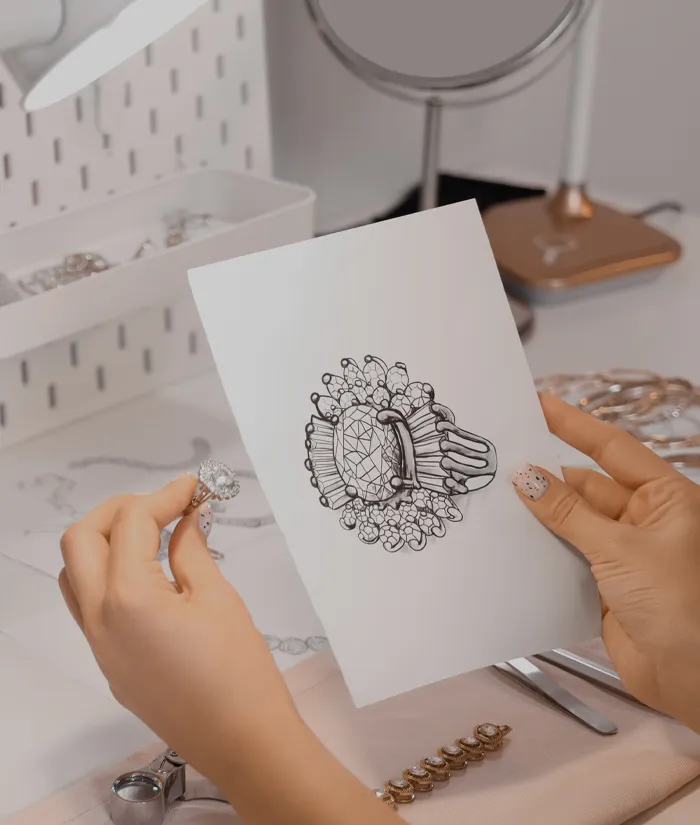 A hand holds a diamond ring next to a sketch of a similar design, showcasing jewelry design at Bryan Jewelry. Start a Project.