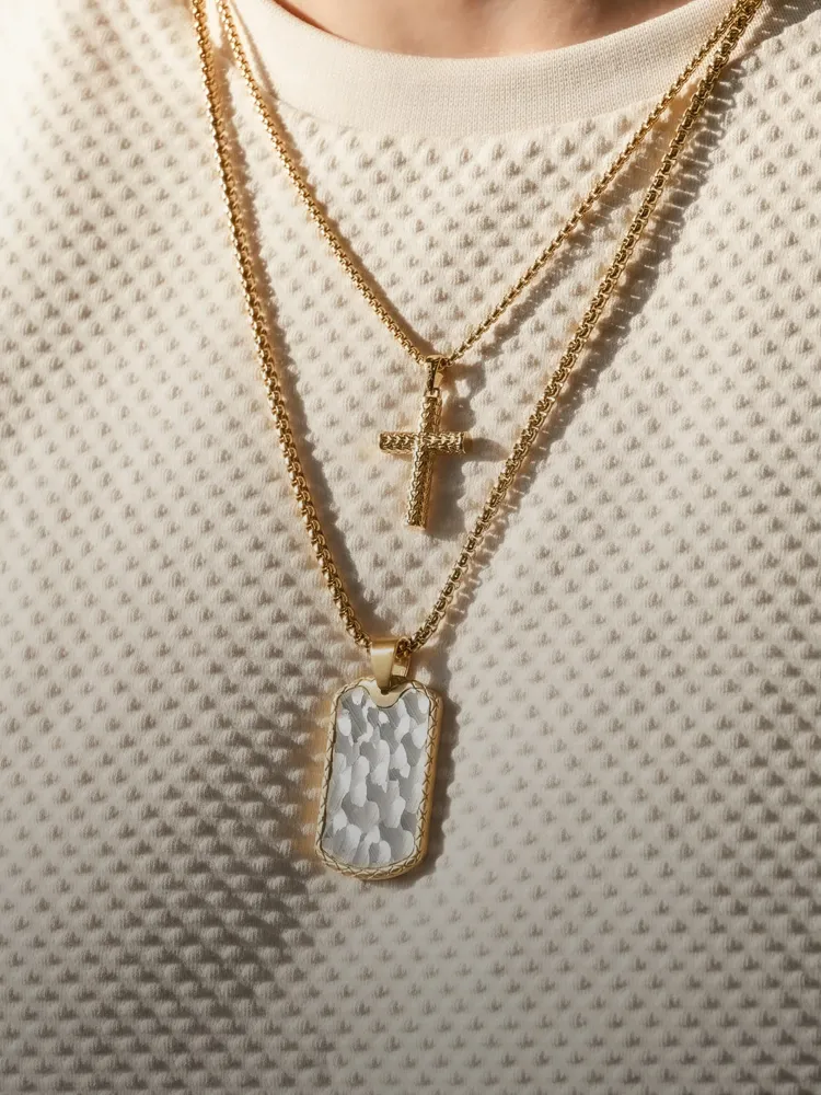 Three gold necklaces: a cross, a textured dog tag, and a chain, worn over a white textured shirt. 