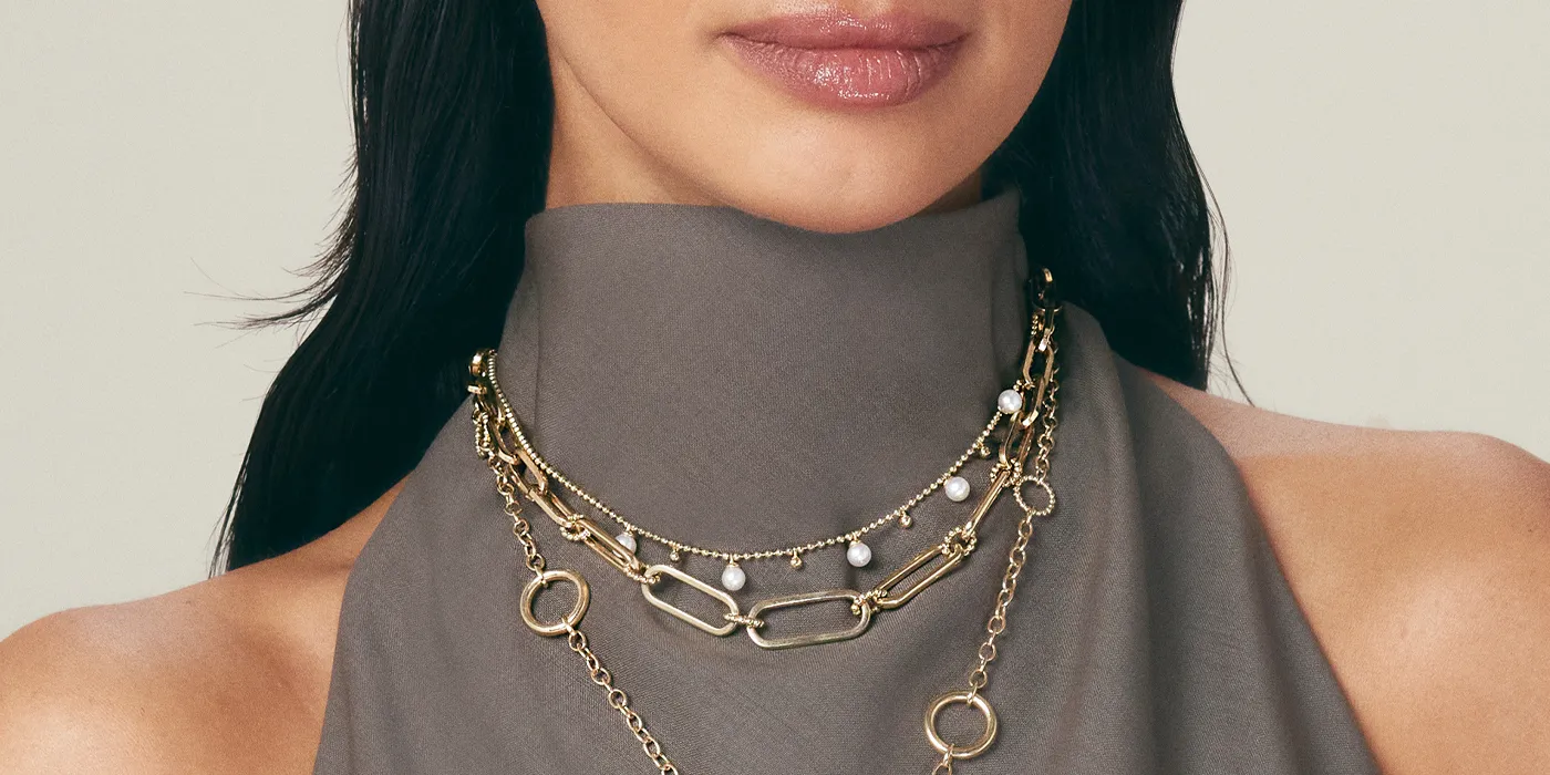 Close-up of layered gold necklaces with pearls, worn by a model in a gray top. Shop at Bryan Jewelry in Prattville.