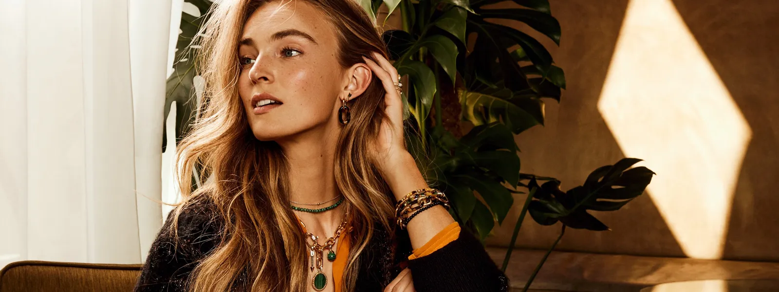 Model showcasing layered jewelry against a backdrop of plants and soft lighting. Visit Bryan Jewelry for fine pieces.
