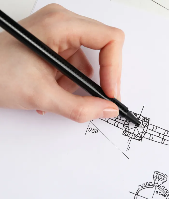 A hand drawing a technical design on paper with a black pen, illustrating custom jewelry design concepts. Learn more.