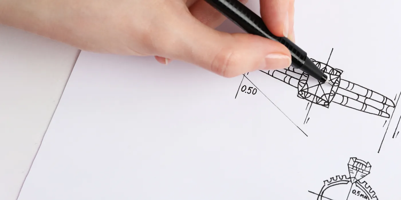 A hand drawing a technical design on paper with a black pen, illustrating custom jewelry design concepts. Learn more.