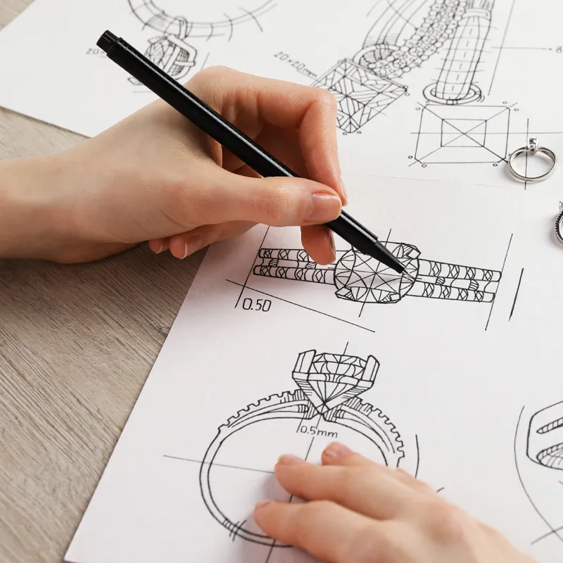 A hand sketches jewelry designs on paper, with two rings placed nearby, showcasing custom jewelry creation at Bryan Jewelry.