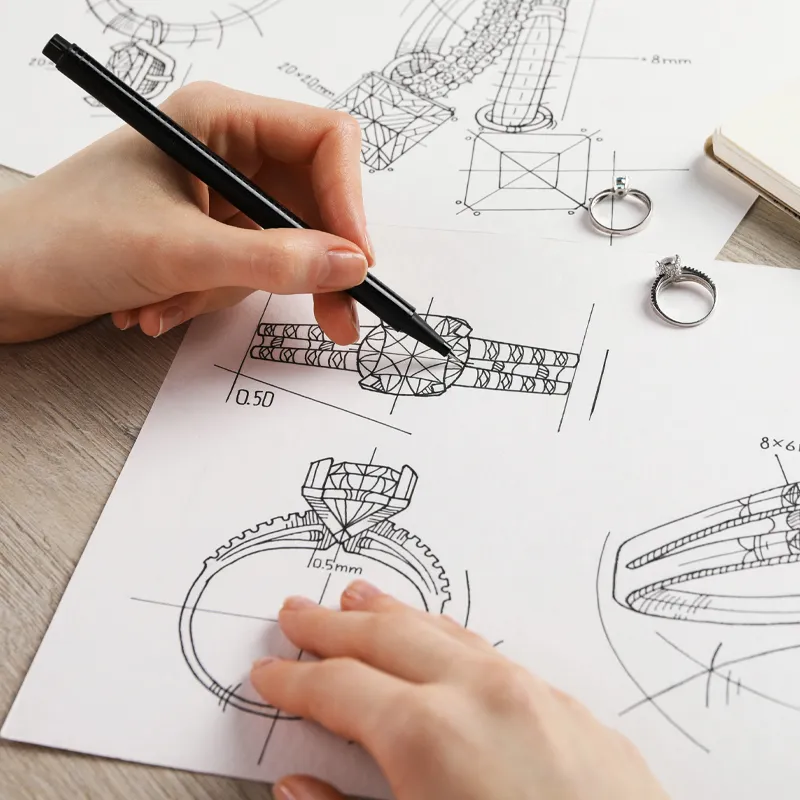 A hand sketches jewelry designs on paper, with two rings placed nearby, showcasing custom jewelry creation at Bryan Jewelry.