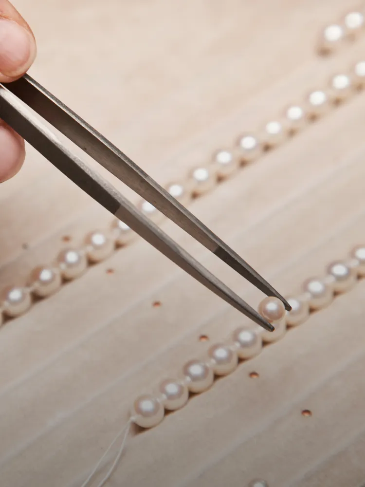 A person uses tweezers to pick up a pearl, surrounded by more pearls on a surface. Click for jewelry repair services.