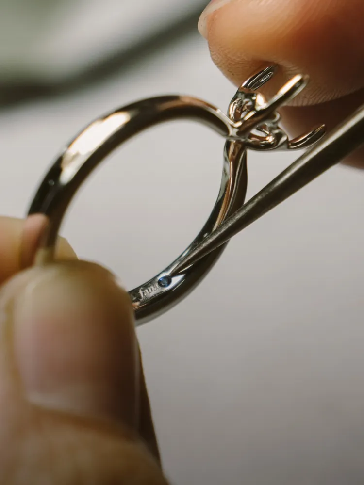 A person inspects a silver ring with tweezers, revealing a small blue stone inside. Click for jewelry repair services.