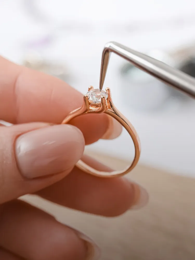 A hand holding a rose gold ring with a diamond, being inspected with tweezers. Click for jewelry repair services.