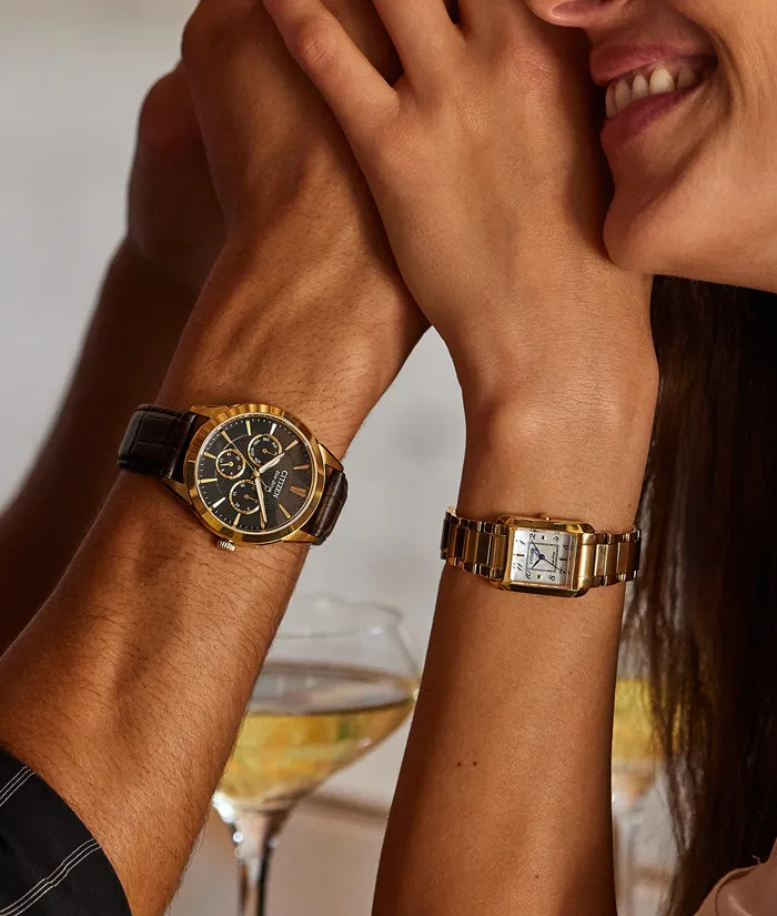 A couple's hands with stylish watches, one gold and black, the other gold and silver, against a blurred background.