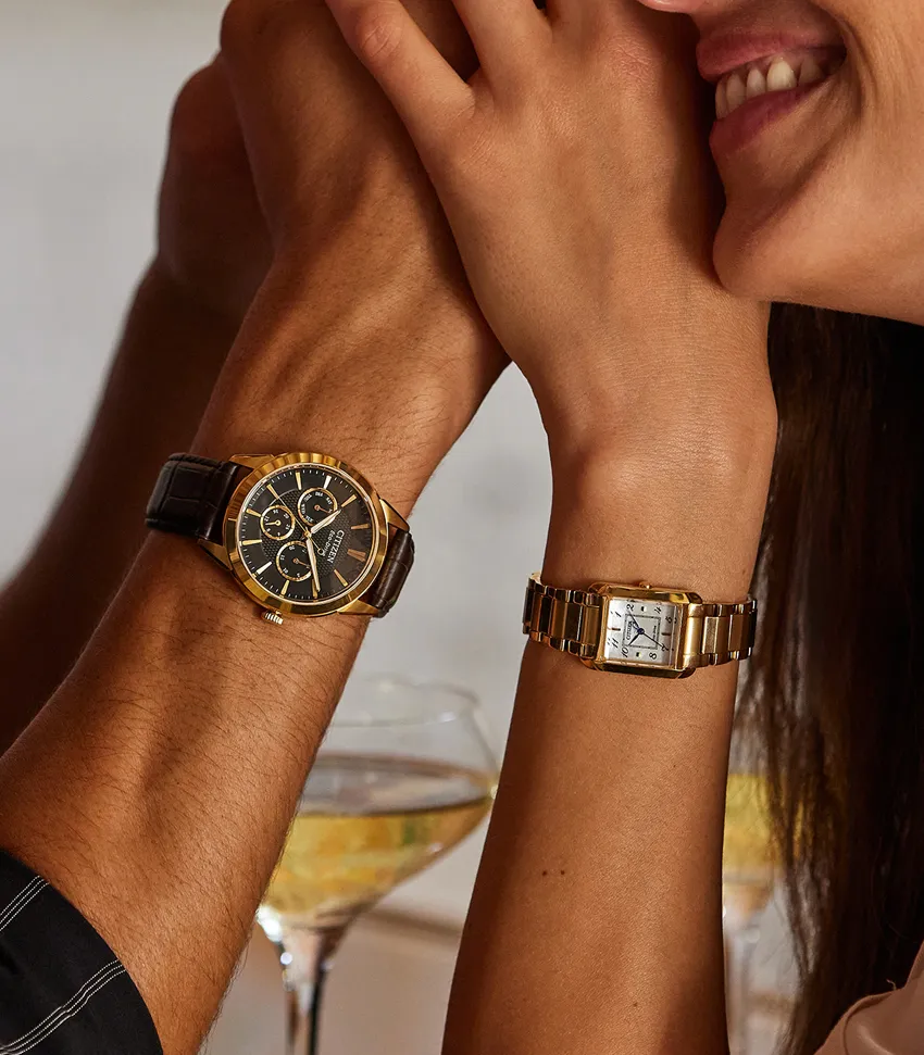 A couple's hands with stylish watches, one gold and black, the other gold and silver, against a blurred background.
