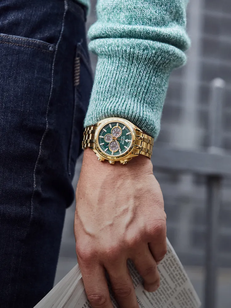 Gold chronograph watch on a man's wrist, holding a newspaper, with a green dial and stylish sweater. Shop at Bryan Jewelry.