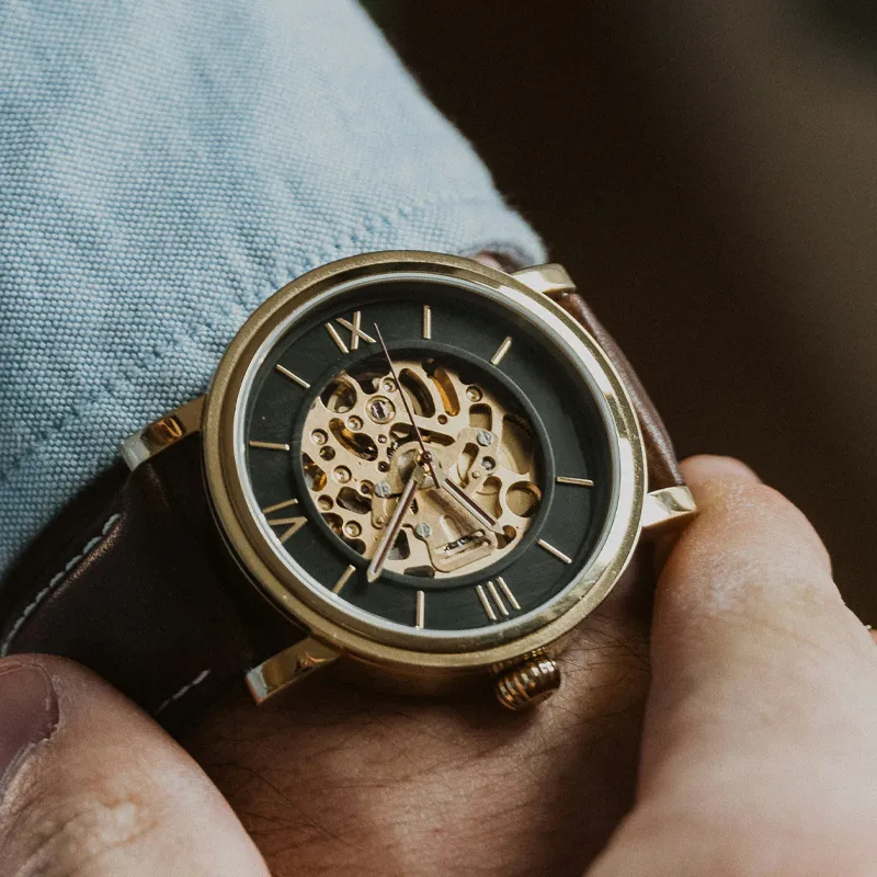 Close-up of a gold skeleton watch on a wrist, featuring a black dial and brown leather strap. Shop at Bryan Jewelry.